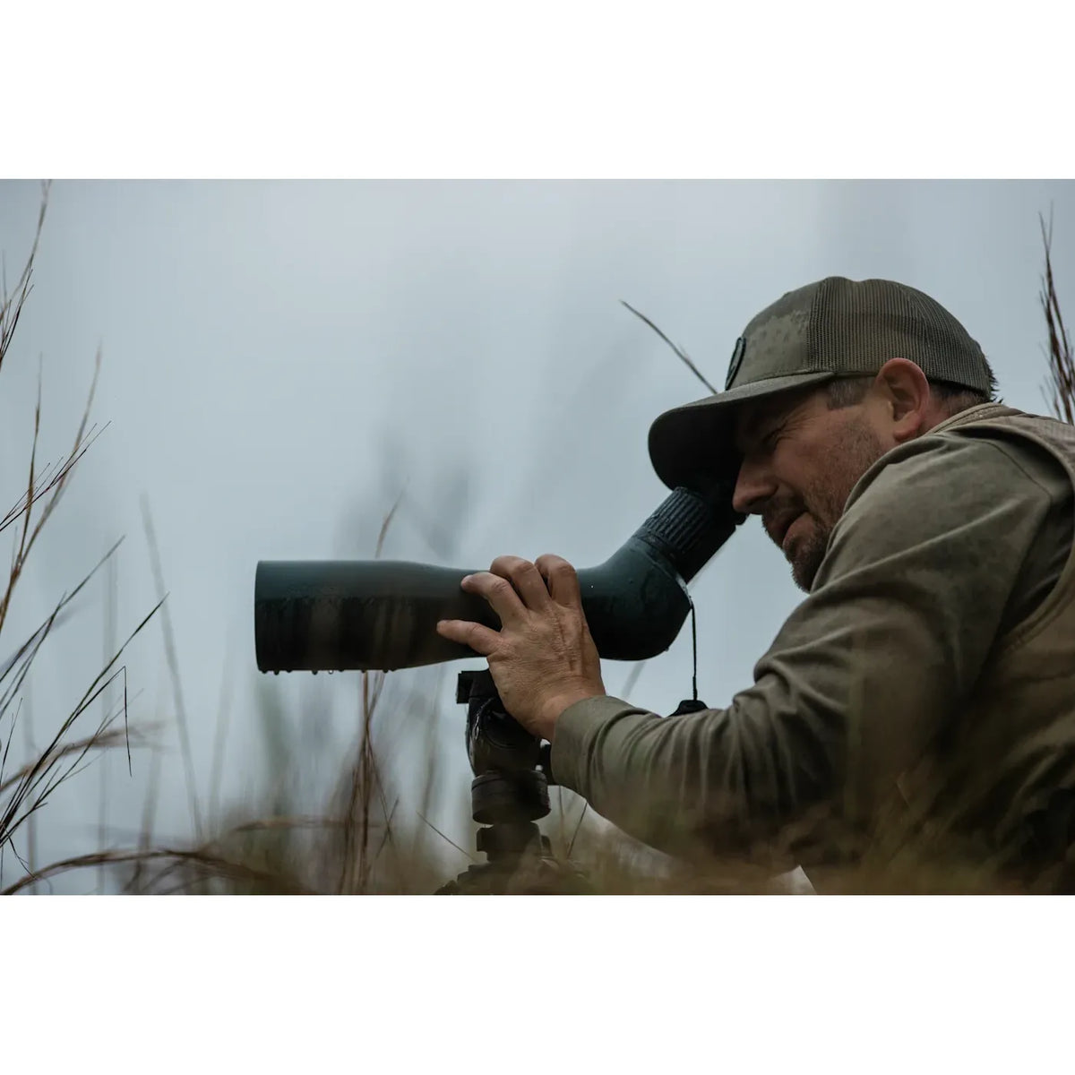 A man in camouflage clothing and a cap looks through a Swarovski Optik AT Endura 21-65x75 spotting scope mounted on a tripod in an outdoor grassy setting. 